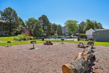A large log sits in the middle of a gravel area with picnic tables and a playground in the background